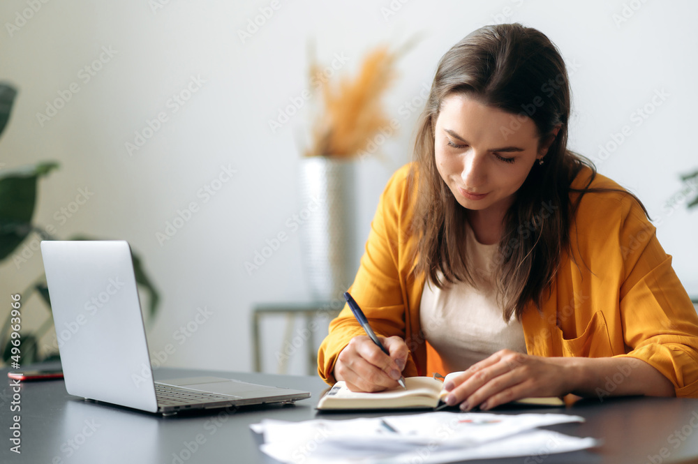 Focused successful smart purposeful young caucasian business woman, manager, student or freelancer, sitting at the table, wearing stylish clothes, using a laptop, taking notes, analyzing, e-learning Focused successful smart purposeful young caucasian business woman, manager, student or freelancer, sitting at the table, wearing stylish clothes, using a laptop, taking notes, analyzing, e-learning
