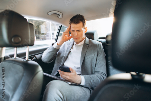 Young businessman tired from work holding hand to his head while sitting in the back seat of the car. © anatoliycherkas