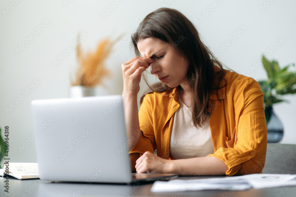 Headache and migraine. Exhausted Caucasian young adult woman, top manager, office worker, tired from work, stressed, took a break from work, needs rest, massages the bridge of her nose, closes eyes Headache and migraine. Exhausted Caucasian young adult woman, top manager, office worker, tired from work, stressed, took a break from work, needs rest, massages the bridge of her nose, closes eyes