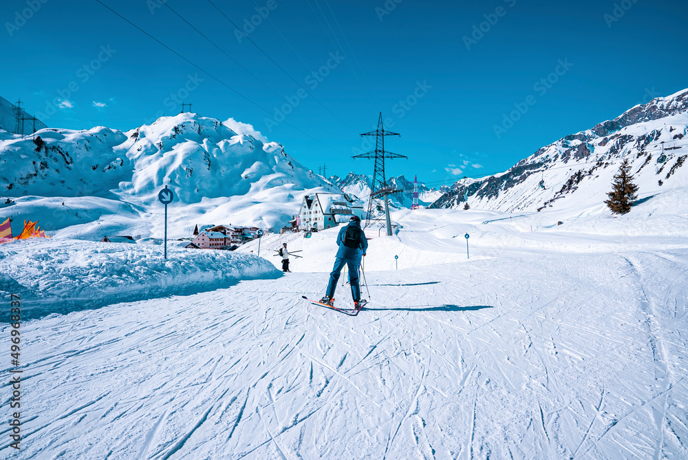 Skier skiing on snowy downhill. Tourist is leading toward houses on mountain against sky. He is enjoying winter sport in alps.
