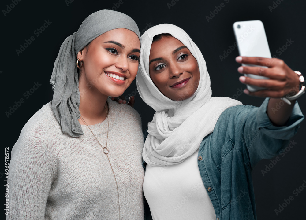 Selfie time. Cropped shot of two attractive young women standing together and wearing hijabs while taking a selfie against a black background.