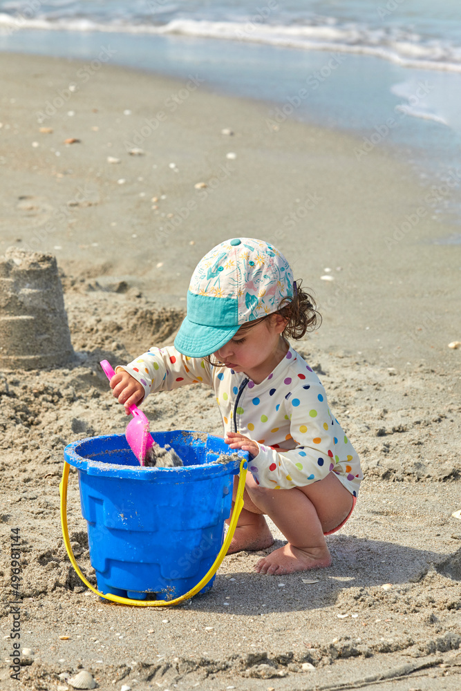 Little girl playing in the sand with a blue bucket and pink shovel making sand castles on a beautiful beach Little girl playing in the sand with a blue bucket and pink shovel making sand castles on a beautiful beach