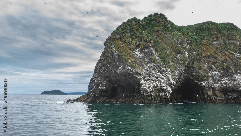 A rocky island in the Pacific Ocean. Seabirds nest on the steep slopes. A dark cave - grotto is visible above the water. Cloudy. Kamchatka. Starichkov Island