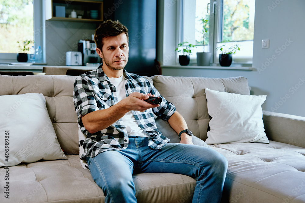 Obraz premium Man sitting on sofa and watching TV, switch channels using remote control. Resting at home, leisure weekend