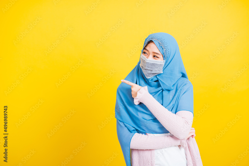 Asian Muslim Arab, Portrait of happy beautiful young woman religious wear veil hijab and face mask protective to prevent coronavirus she pointing finger side away space, isolated on yellow background Asian Muslim Arab, Portrait of happy beautiful young woman religious wear veil hijab and face mask protective to prevent coronavirus she pointing finger side away space, isolated on yellow background