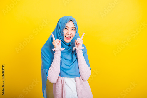 Asian Muslim Arab, Portrait of happy beautiful young woman Islam religious wear veil hijab funny smile she positive expression pointing with finger up to space isolated yellow background