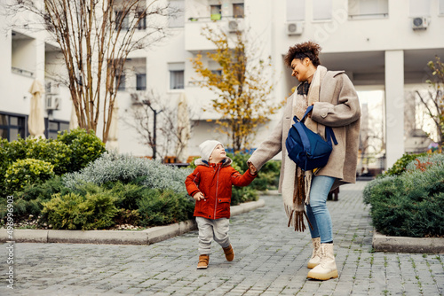 A playful babysitter holding hands with the little boy and taking him to the daycare or pre-school.