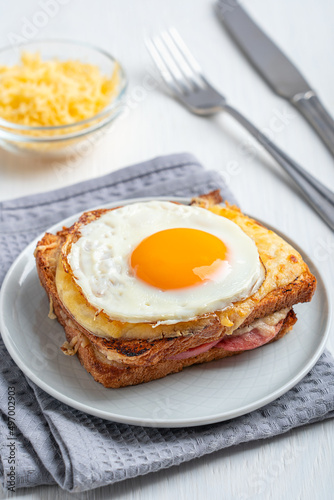French traditional Croque madame baked sandwich made of toasted bread, ham and cheese with fried egg on top served on plate with towel and tableware as snack for breakfast on white wooden table