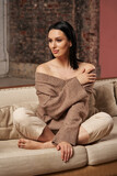 a beautiful woman with dark hair with a smooth hairstyle gracefully sits on a sofa in cozy clothes, with bare shoulders