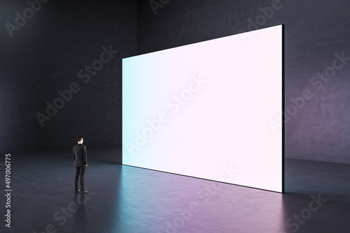 Tiny young european businessman looking at huge white mock up billboard in dark concrete interior.