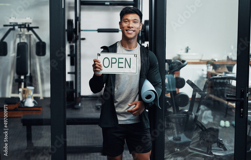 Youre welcome any time between 9 and 5. Cropped portrait of a handsome young male fitness instructor holding up a sign that says OPEN while standing in a gym.