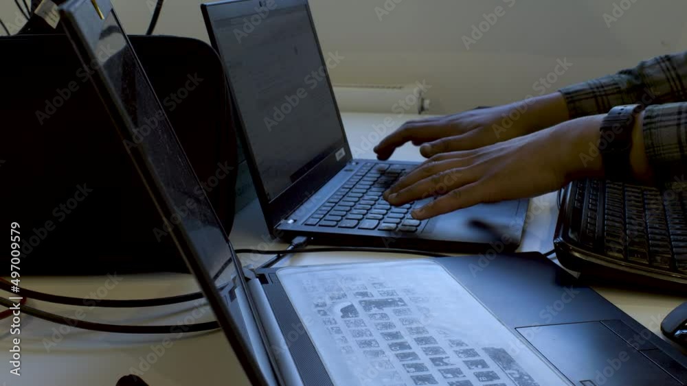 A close up shot of a busy and cluttered work desk of an IT professional as he transfers data from one laptop to another while continuing to type on an external keyboard