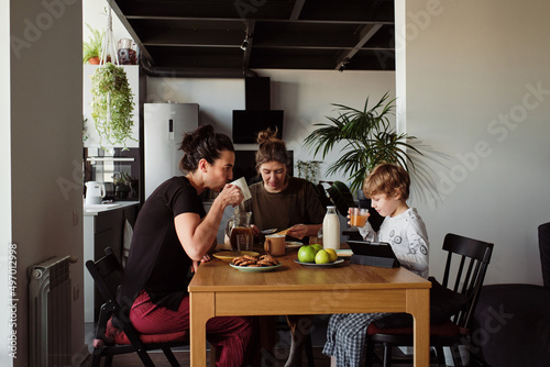 Family of two young women with little boy sitting at table and having breakfast in kitchen
