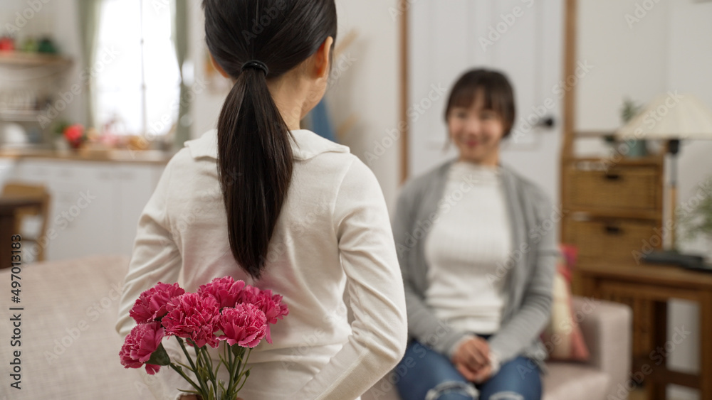 rear view of asian little girl giving mother carnation bouquet on mother's day at home. she hides flower on back and walks up to her smiling mom