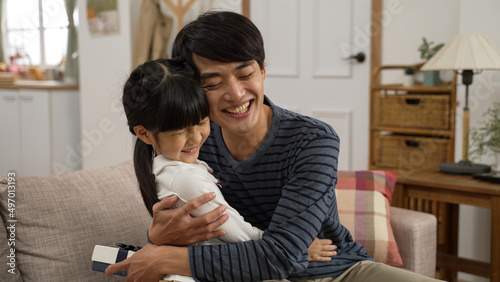 Asian girl walking towards dad and showing him surprise gift on Father’s Day at home. the touched man giving his daughter a big hug on the sofa