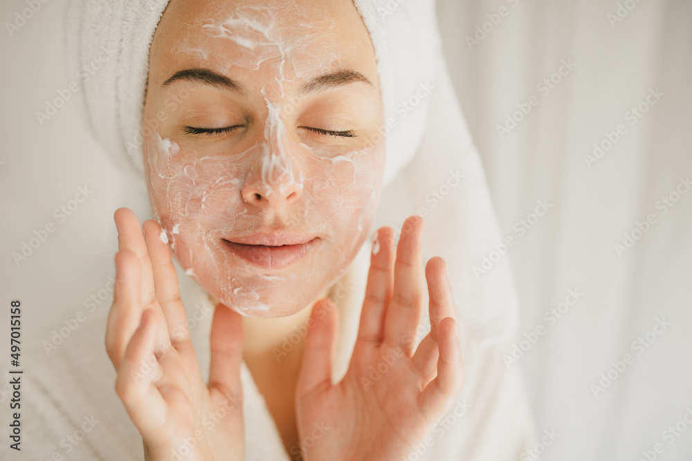 Young beautiful woman wearing bathrobe and towel on her hair applying moistrizing cream on her face. Skin care morning rituals. Beauty routine.