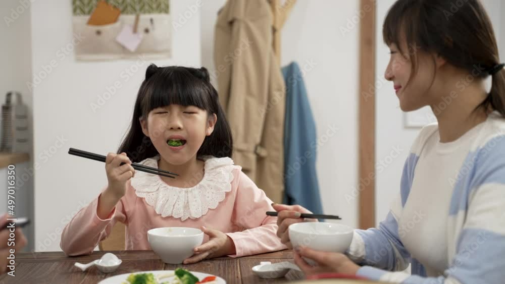 closeup of cute asian girl enjoying eating healthy green vegetable with chopsticks at dining table. she smiles at her mother as she is chewing the food closeup of cute asian girl enjoying eating healthy green vegetable with chopsticks at dining table. she smiles at her mother as she is chewing the food