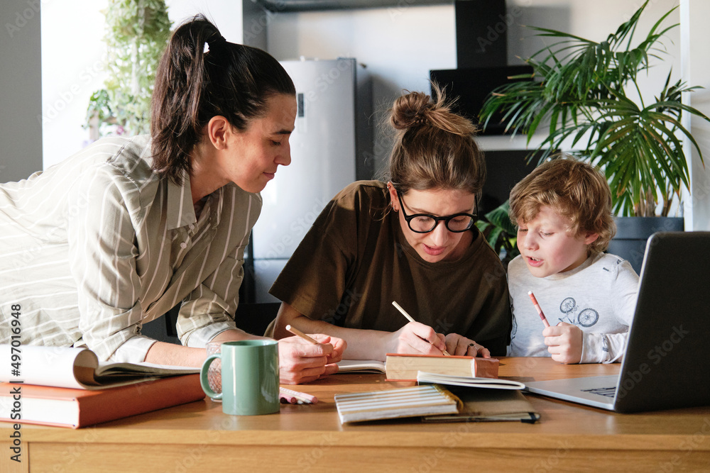 Homosexual mothers sitting at table with books and helping their son with homework Homosexual mothers sitting at table with books and helping their son with homework