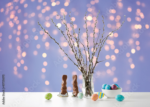 easter, holidays and object concept - pussy willow branches, colored eggs and chocolate bunnies on table over festive lights on violet background photo