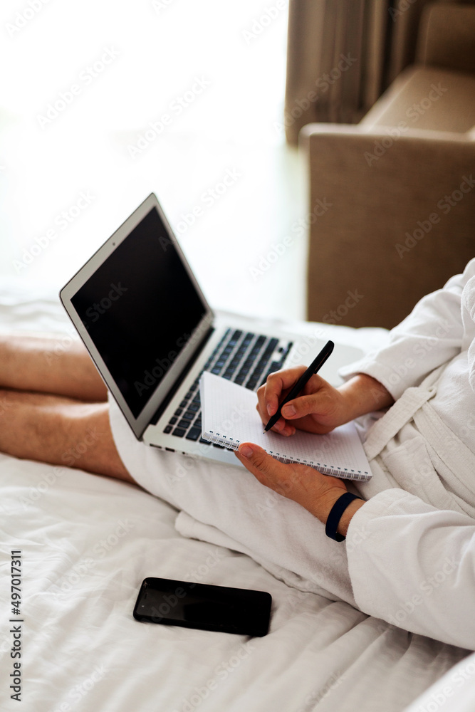 a man in a bathrobe with a laptop on his lap lies in the bed and writes something in a notebook a man in a bathrobe with a laptop on his lap lies in the bed and writes something in a notebook