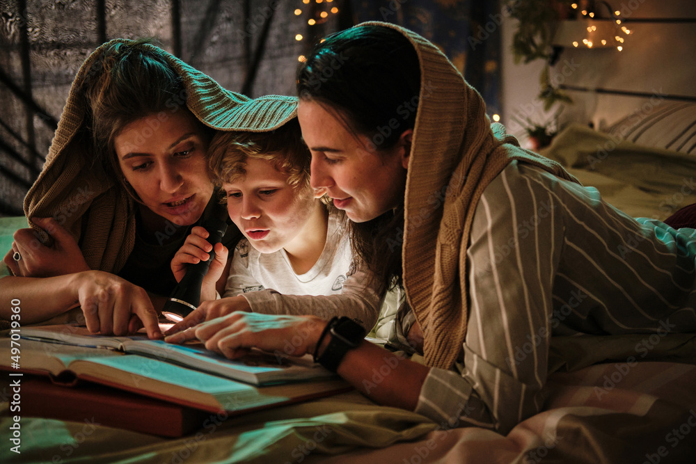 Homosexual mothers lying on bed under blanket with their son and reading a book together using flashlight