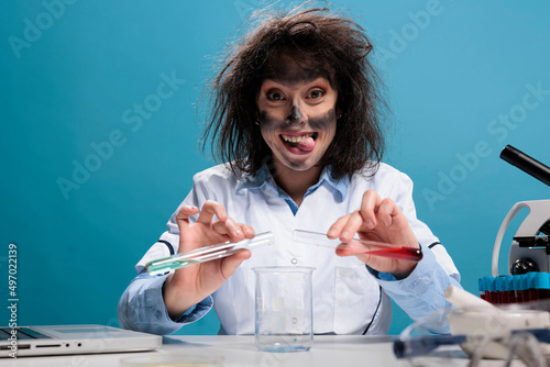 Lunatic goofy chemist with wild face expression testing new chemical formula in laboratory space on blue background. Crazy lab worker grinning dreadful at camera while mixing chemical compounds. photo