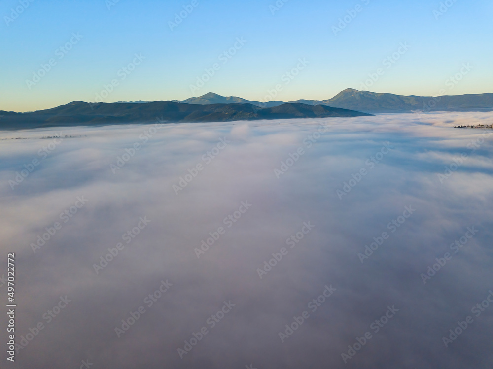 Flight over fog in Ukrainian Carpathians in summer. Mountains on the horizon. A thick layer of fog covers the mountains with a continuous carpet. Aerial drone view.