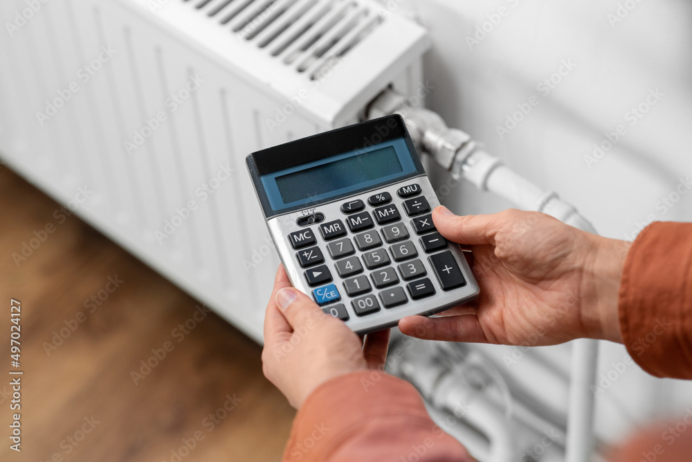 heating, energy crisis and consumption concept - close up of hand with calculator and radiator at home