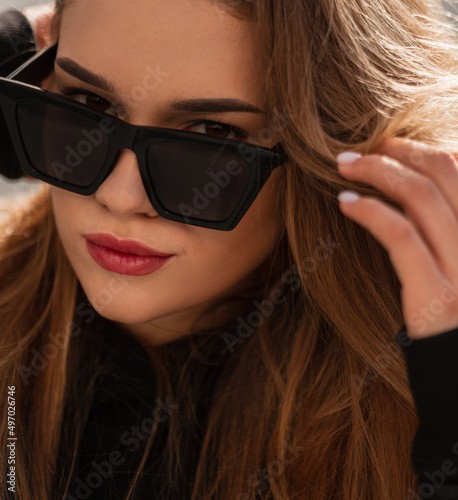 Stylish portrait of a beautiful fashionable woman with trendy sunglasses in a black trendy vintage leather jacket and hoodie looking at the camera