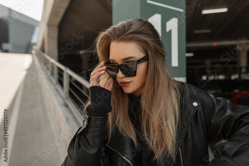 Urban fashionable portrait of beautiful girl model with rock stylish leather jacket and black hoodie wear a trendy sunglasses in the city