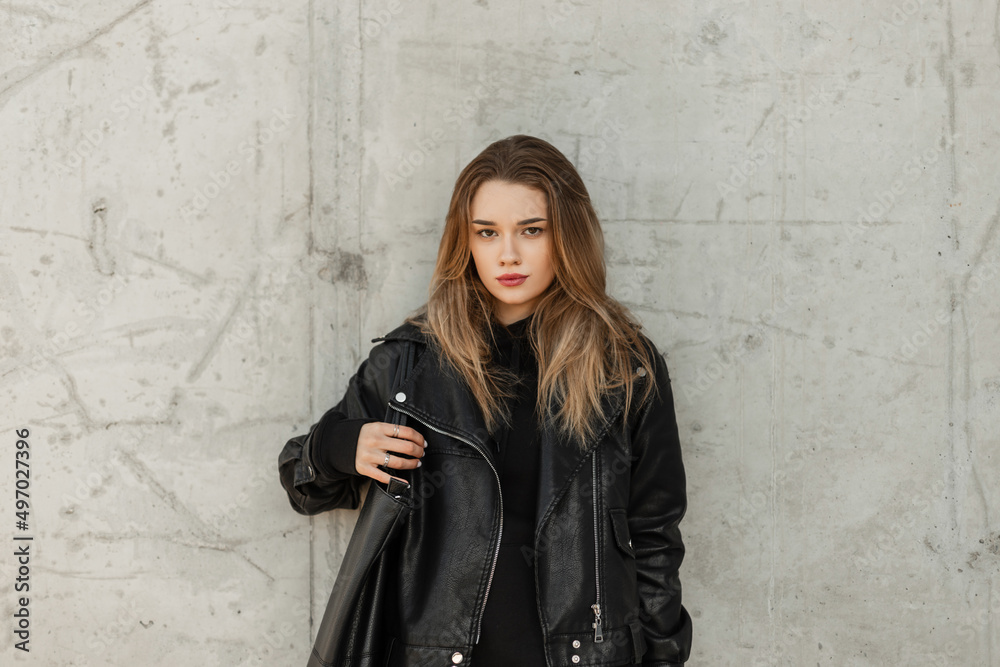 Fashionable adorable woman model with fashion black leather jacket and black hoodie with leather bag stands near a gray concrete wall