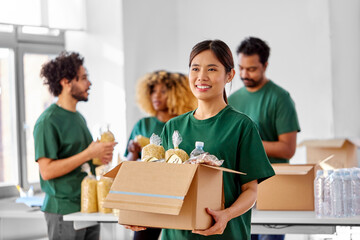 charity, donation and volunteering concept - happy smiling female volunteer with food in box and international group of people at distribution or refugee assistance center