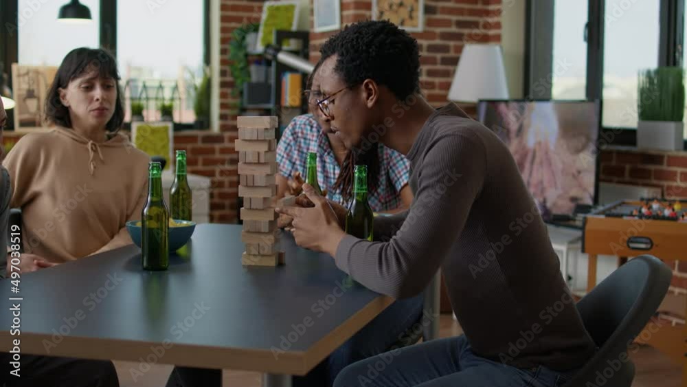Men and women gathering at home to play jenga game with wooden blocks and square tower building on table. Group of friends playing competitive board games to have fun with construction.