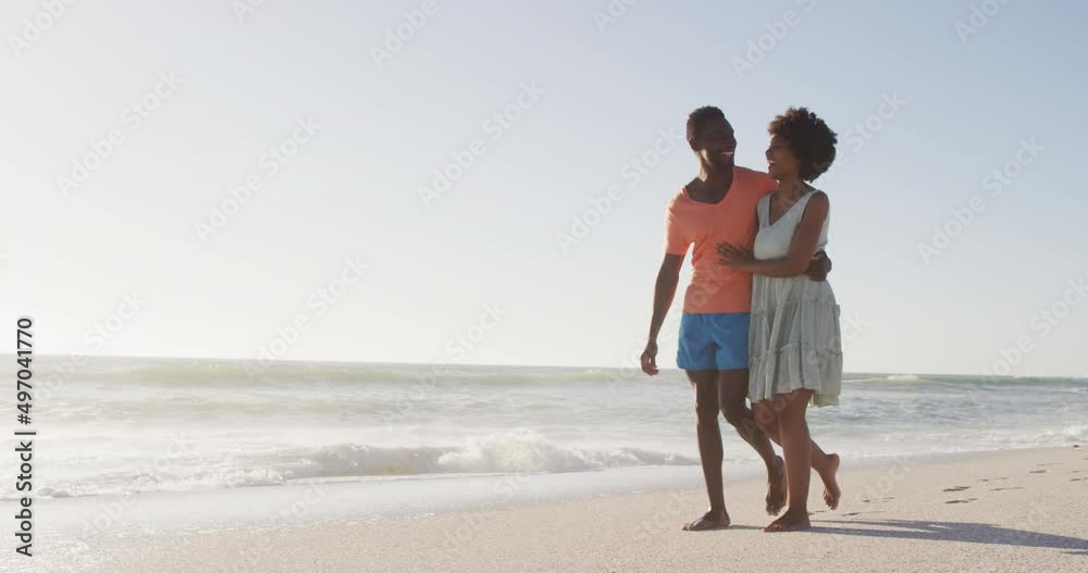 Smiling african american couple embracing and walking on sunny beach Smiling african american couple embracing and walking on sunny beach