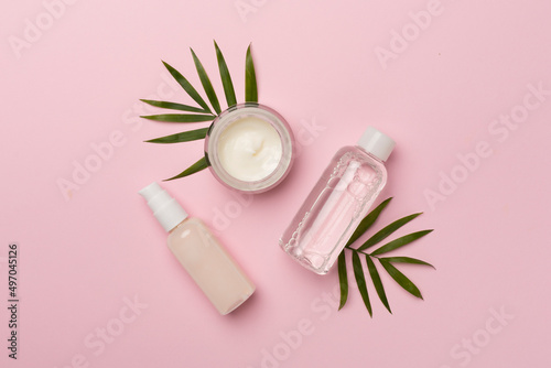 Facial tonic and cream with green leaves on color background, top view