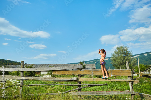 A school-age girl stands on a wooden rustic fence surrounded by wild grasses and flowers. © Fotoproff