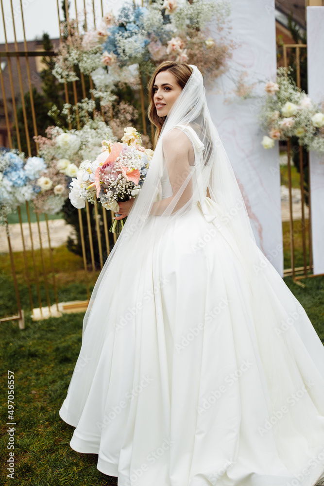 Fototapeta premium Full length portrait of attractive young bride in a white wedding dress and veil holding a big bouquet of white and pink flowers in her hands on a background of wedding ceremony arch