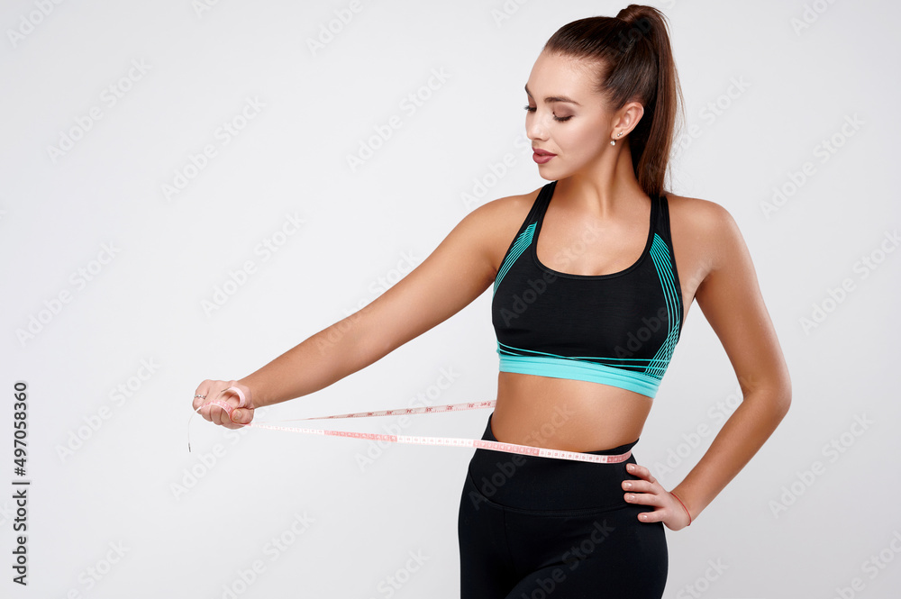 Strong slim attractive white fitness female in sport suit measuring waist on gray isolated background Strong slim attractive white fitness female in sport suit measuring waist on gray isolated background