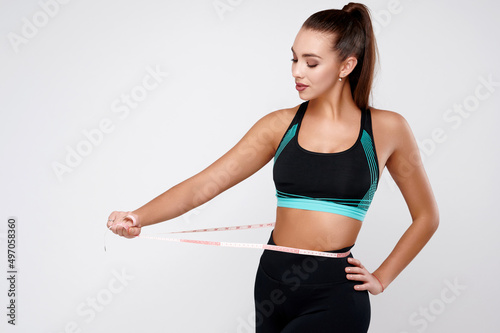 Strong slim attractive white fitness female in sport suit measuring waist on gray isolated background © khmelev