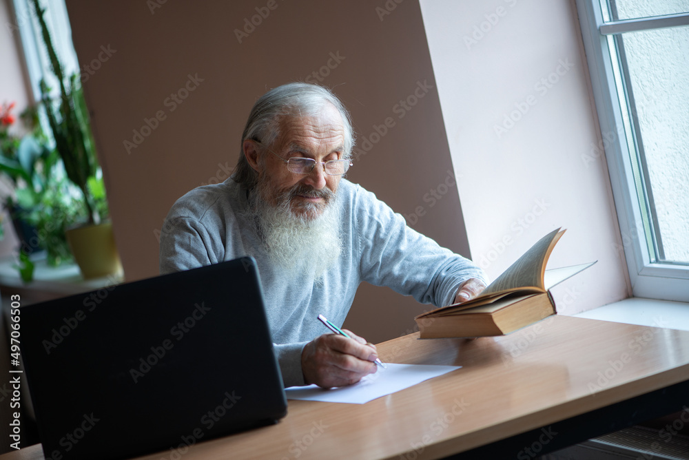 Bearded old man with laptop and book sitting at table and write notes Bearded old man with laptop and book sitting at table and write notes