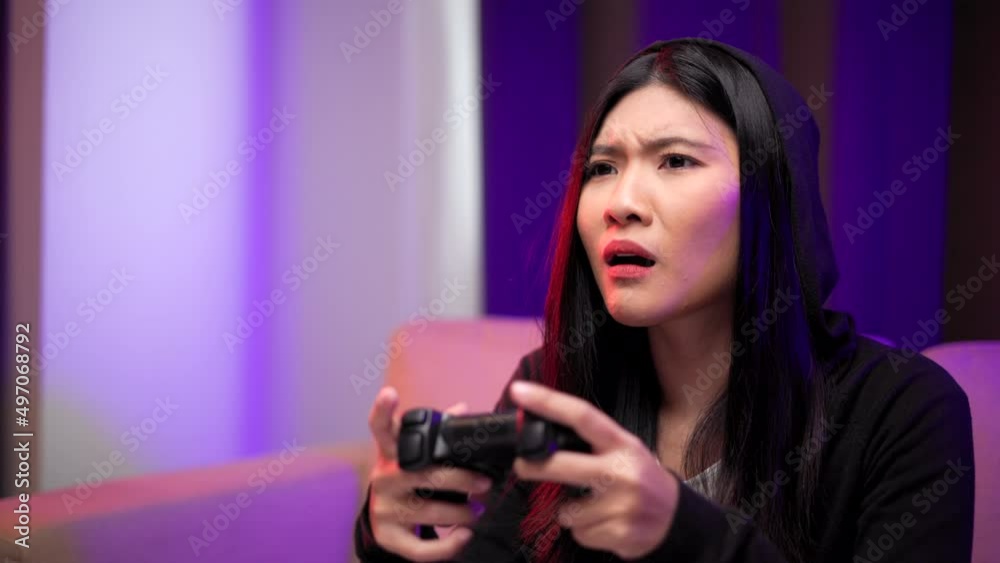 Gamer losing moment. Young asian pretty woman sitting on sofa holding joystick in living room. Unhappy female Professional Streamer chinese playing game online in dark room neon light. Gamer losing moment. Young asian pretty woman sitting on sofa holding joystick in living room. Unhappy female Professional Streamer chinese playing game online in dark room neon light.