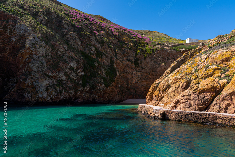 idyllic cove with small beach on a small Atlantic island wirth cliffs and many colorful flowers under a blue sky