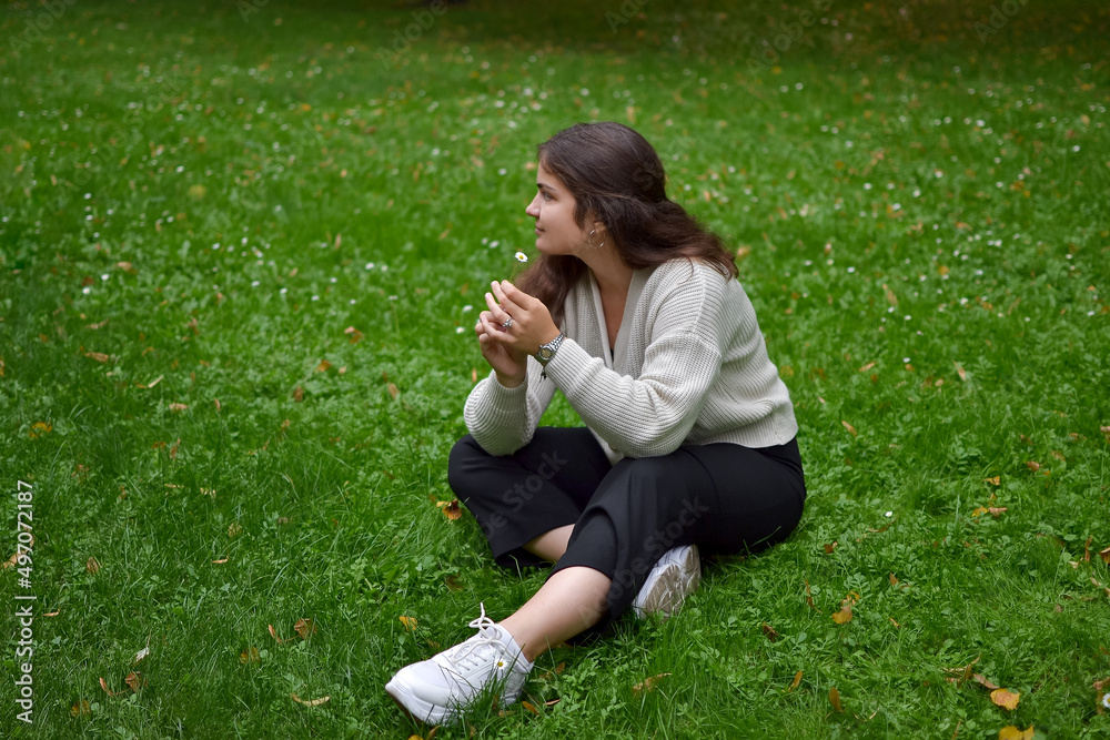 Outdoors, a woman in a beige knitted sweater and trousers sits on the grass in the park, holding a chamomile flower in her hand and looking away, resting and recuperating after a hard day's work. Outdoors, a woman in a beige knitted sweater and trousers sits on the grass in the park, holding a chamomile flower in her hand and looking away, resting and recuperating after a hard day's work.