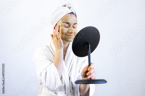 Tableau sur toile Young woman with a green mask on her face in a bathrobe in front of a mirror