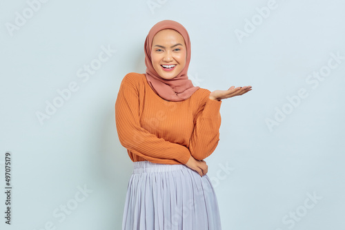 Smiling beautiful asian woman in brown sweater and hijab pointing to copy space with palms, showing advertising products and confidently looking at camera isolated over white background. © Bangun Stock Photo