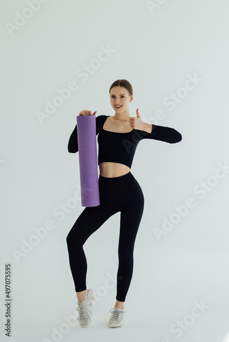 Beautiful smiling young woman in sportswear holding a yoga mat giving thumbs up, isolated on white background. Healthy lifestyle concept. Fitness and yoga concept.