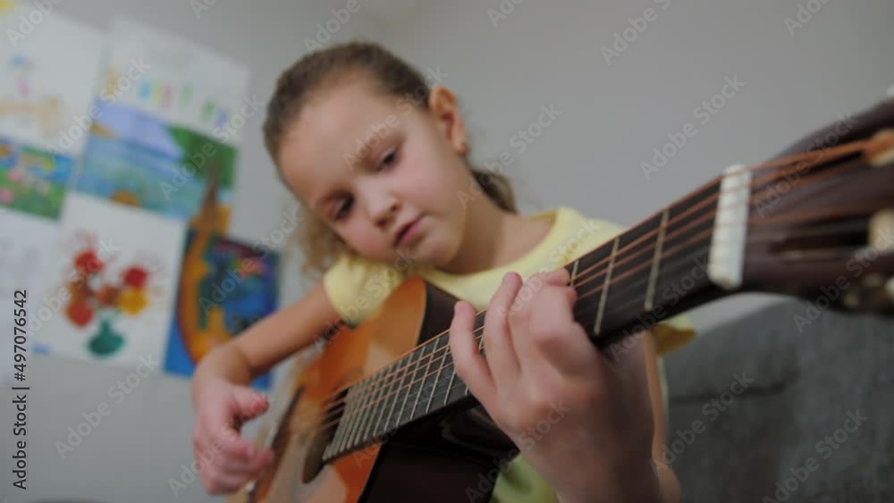 Little caucasian girl, school age child playing acoustic guitar sitting on sofa. Hobby, learning new skill, musical instrument practice, spending free time at home 