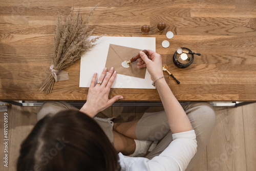 Top view of unrecognizable woman making white wax seal with stamp on brown greeting envelope, sitting at wooden desk.