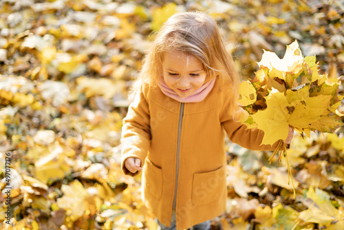 A happy little girl in a coat walks in the park in autumn through the fallen yellow foliage and rejoices.