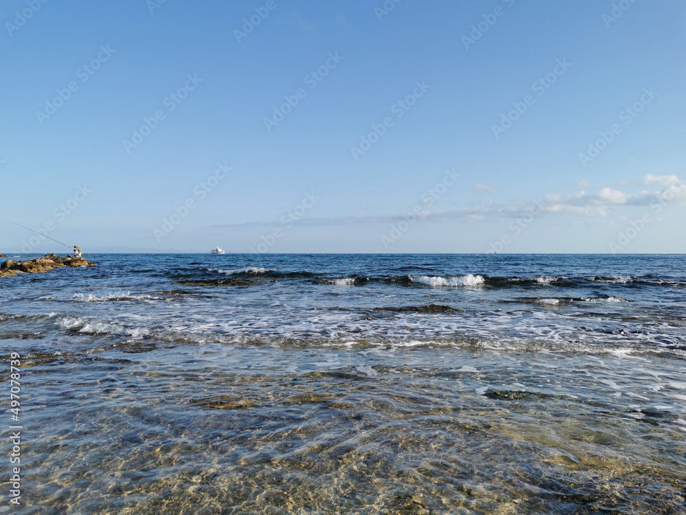 The coast of the Mediterranean Sea, a stone ridge, at the end of which is a fisherman, in the sea a white pleasure yacht against a blue sky with clouds. The coast of the Mediterranean Sea, a stone ridge, at the end of which is a fisherman, in the sea a white pleasure yacht against a blue sky with clouds.